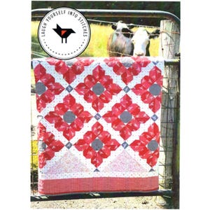 May include: A red, white, and black quilt with a floral pattern hangs on a fence. The quilt is made with a variety of fabrics, including gingham and polka dots. The quilt is hanging in front of a green field with a cow in the background. The quilt has the text "Laugh Yourself Into Stitches" on a black circle with a red bird.