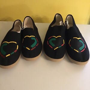 May include: Pair of black canvas slip-on shoes with a heart design in red, yellow, and green.