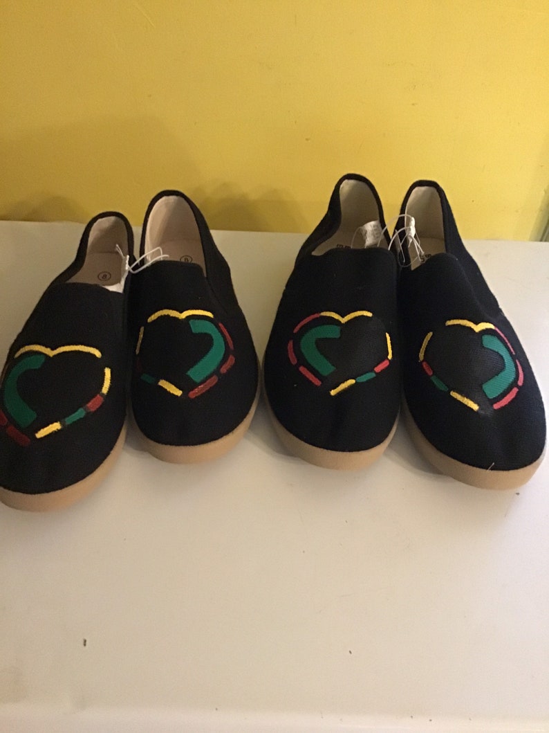 May include: Pair of black canvas slip-on shoes with a heart design in red, yellow, and green.