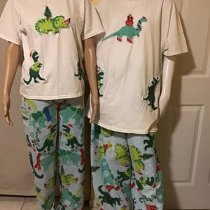 Puede incluir: Dos camisetas blancas con aplicaciones de dinosaurios verdes, una con un &aacute;rbol de Navidad en la parte superior del dinosaurio. Pantalones de pijama a juego azul y verde con un patr&oacute;n de dinosaurio y &aacute;rbol de Navidad.