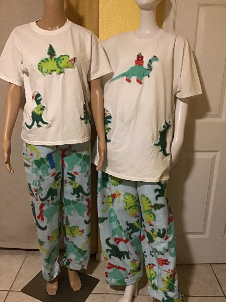 Puede incluir: Dos camisetas blancas con aplicaciones de dinosaurios verdes, una con un &aacute;rbol de Navidad en la parte superior del dinosaurio. Pantalones de pijama a juego azul y verde con un patr&oacute;n de dinosaurio y &aacute;rbol de Navidad.