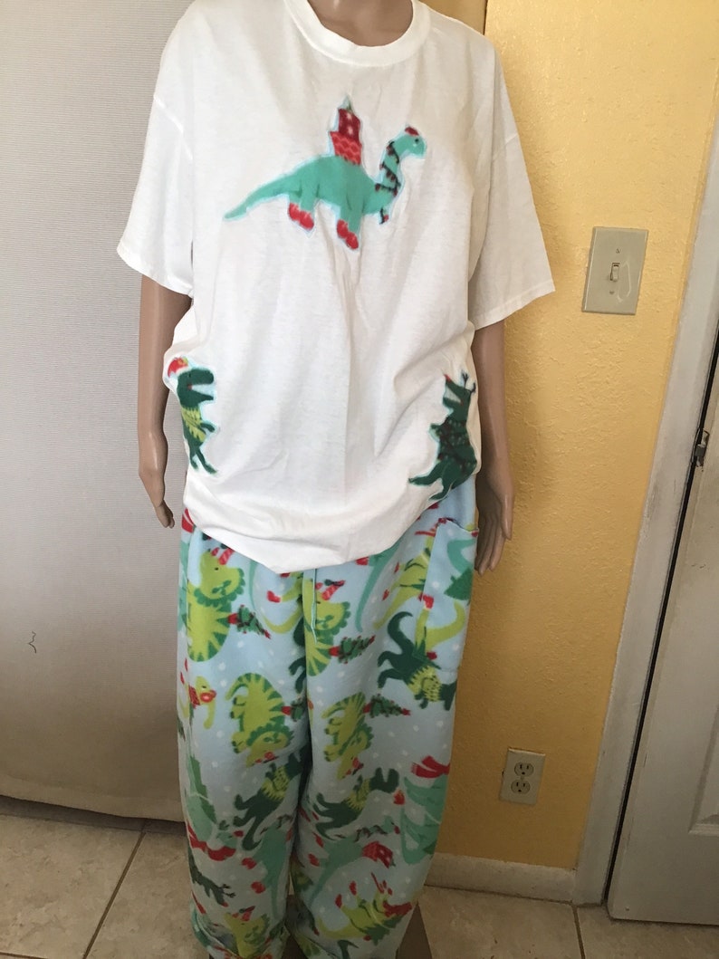 Puede incluir: Camiseta blanca con un dinosaurio verde que lleva un gorro de Pap&aacute; Noel rojo y un &aacute;rbol de Navidad verde y rojo en la parte delantera. Pantal&oacute;n de pijama a juego con estampado de dinosaurio verde y rojo.