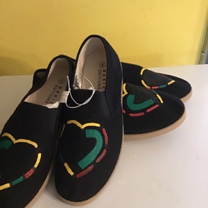 May include: Two pairs of black canvas slip-on shoes with a red, yellow, and green design. The shoes have the text "BASIC EDITIONS" on the side.