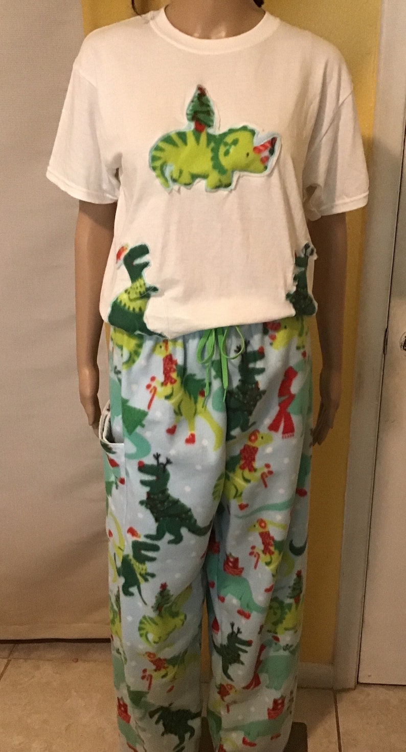 Puede incluir: Camiseta blanca con un dinosaurio verde que lleva un gorro de Pap&aacute; Noel y un &aacute;rbol de Navidad en la espalda. Pantal&oacute;n de pijama azul a juego con estampado de dinosaurio verde y &aacute;rboles de Navidad.
