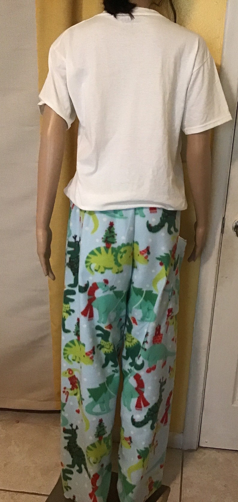Puede incluir: Pantal&oacute;n de pijama azul con estampado de dinosaurio navide&ntilde;o. Los dinosaurios llevan gorros de Pap&aacute; Noel y bufandas. El pantal&oacute;n tiene un fondo blanco con lunares blancos.