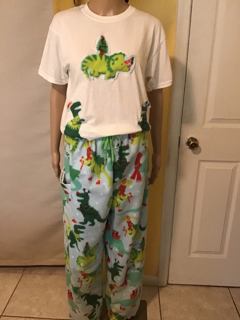 Puede incluir: Camiseta blanca con un dinosaurio verde que lleva un gorro de Pap&aacute; Noel y sostiene una aplicaci&oacute;n de &aacute;rbol de Navidad. Pantal&oacute;n de pijama azul a juego con estampado de dinosaurio verde.
