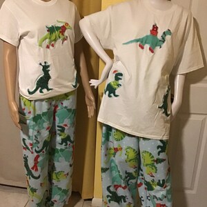 Puede incluir: Camiseta blanca con un dinosaurio verde que lleva un gorro de Pap&aacute; Noel y una bufanda roja. El dinosaurio sostiene un &aacute;rbol de Navidad. La camiseta se combina con pantalones de pijama azules con estampado de dinosaurio verde.