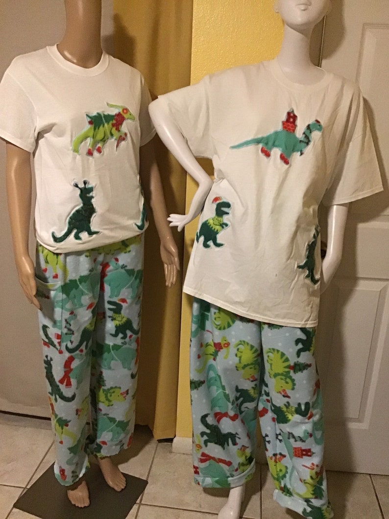 Puede incluir: Camiseta blanca con un dinosaurio verde que lleva un gorro de Pap&aacute; Noel y una bufanda roja. El dinosaurio sostiene un &aacute;rbol de Navidad. La camiseta se combina con pantalones de pijama azules con estampado de dinosaurio verde.