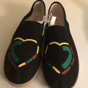 May include: A pair of black canvas slip-on shoes with a heart design. The heart is made up of red, yellow, and green lines.