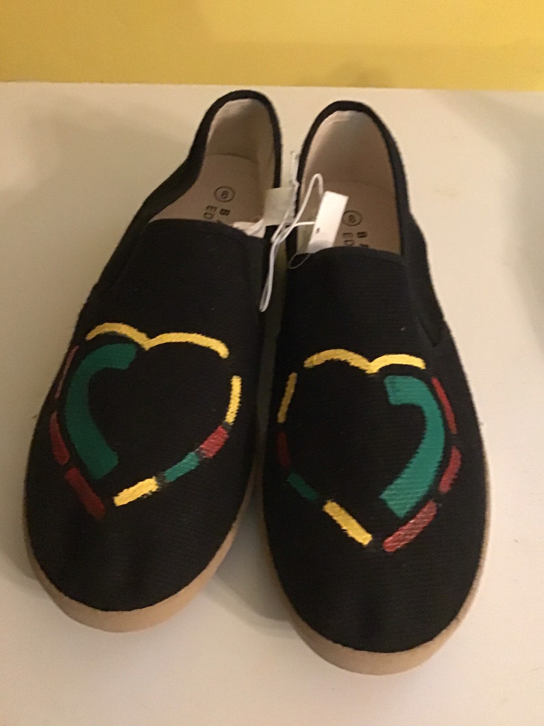 May include: A pair of black canvas slip-on shoes with a heart design. The heart is made up of red, yellow, and green lines.