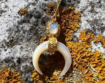 May include: A gold-tone necklace featuring a crescent-shaped white stone pendant suspended from a delicate chain. The pendant includes an oval-shaped iridescent stone set in a gold-tone frame with intricate metalwork details above the crescent.