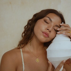 May include: A woman wearing gold jewelry, including a necklace with a sun pendant and a ring. She is holding a large, white, conch shell. The background is a light beige color. The woman has long, wavy brown hair and is wearing a patterned top.