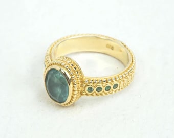 May include: A gold ring featuring an oval, teal-colored gemstone. The ring has a textured band and is accented with small green gemstones. The ring is set against a white background.