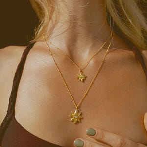 以下が含まれることがあります： 星型のペンダントが付いた2つのゴールドネックレス。ネックレスは重ね付けされており、片方のペンダントはもう一方よりもわずかに小さくなっています。ジュエリーは濃いバーガンディ色のトップスの上から着用されています。グリーンのマニキュアとゴールドの指輪をした人の手が写っています。