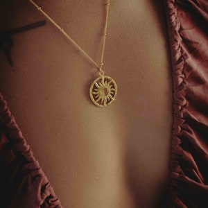 May include: A gold-colored necklace featuring a sun pendant. The pendant is circular with a detailed sun design. The necklace is worn against a dark red top.