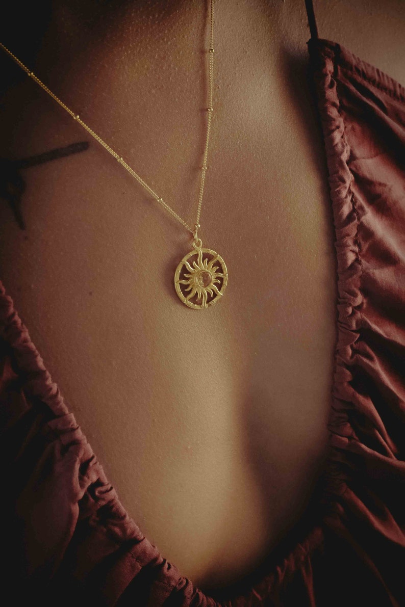 May include: A gold-colored necklace featuring a sun pendant. The pendant is circular with a detailed sun design. The necklace is worn against a dark red top.