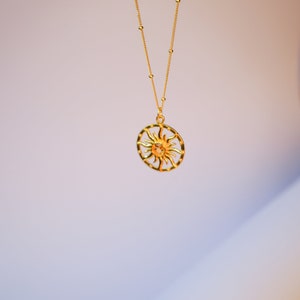 May include: A gold-colored necklace with a sun pendant. The pendant features a sun design within a circular frame. The necklace chain is also gold and has small, evenly spaced beads. The background is a gradient of white and purple.