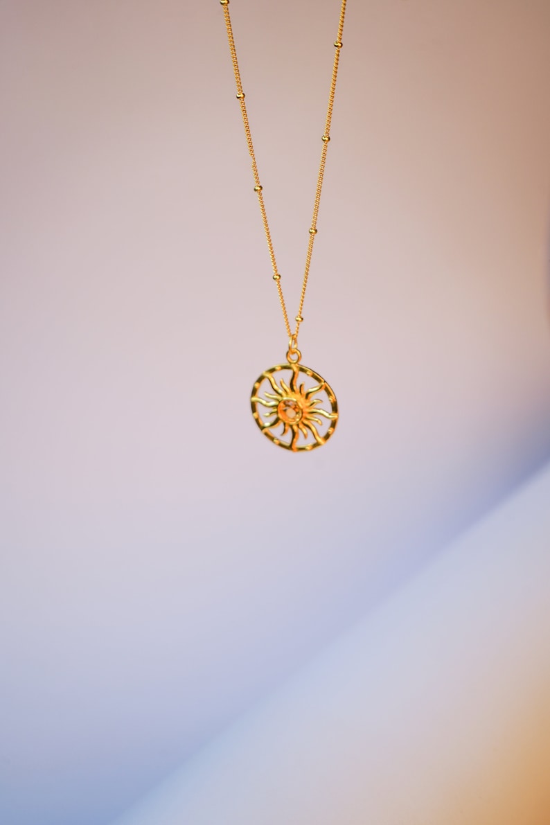 May include: A gold-colored necklace with a sun pendant. The pendant features a sun design within a circular frame. The necklace chain is also gold and has small, evenly spaced beads. The background is a gradient of white and purple.