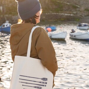May include: A woman wearing a blue beanie with a pom pom and a brown jacket is carrying a white tote bag with a design of five oars and the text "LINO LOKS".