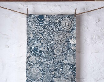 May include: A navy blue tea towel with a white illustrated pattern of seashells and marine life. The towel is hanging from a clothesline, held by a wooden peg. The background features a white wall and a wooden beam. The design is detailed and artistic.
