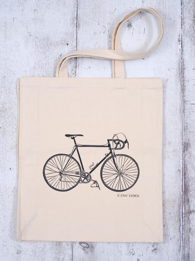 May include: A white canvas tote bag with a black silhouette of a bicycle printed on the front. The text "LINO LORD" is printed below the bicycle.