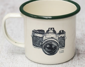 May include: White enamel mug with a green rim featuring a black and white illustration of a vintage camera. The text "L'INO LORD" is printed below the camera.