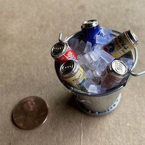 Puede incluir: Un peque&ntilde;o cubo de plata lleno de hielo y cuatro latas de refresco en miniatura. Las latas est&aacute;n etiquetadas con "Coca-Cola", "Coors" y "Budweiser".
