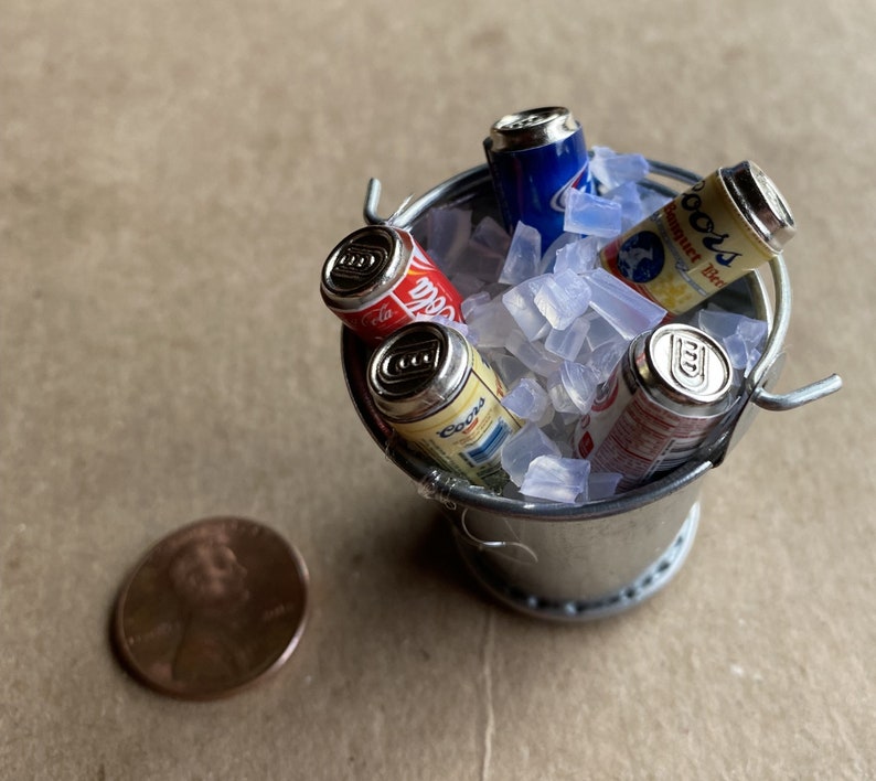 Puede incluir: Un peque&ntilde;o cubo de plata lleno de hielo y cuatro latas de refresco en miniatura. Las latas est&aacute;n etiquetadas con "Coca-Cola", "Coors" y "Budweiser".