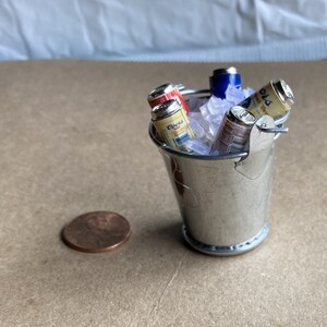 Puede incluir: Un cubo de metal plateado en miniatura lleno de hielo y latas de cerveza en miniatura. Las latas tienen etiquetas con las palabras "Budweiser" y "Corona".