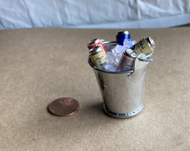 Puede incluir: Un cubo de metal plateado en miniatura lleno de hielo y latas de cerveza en miniatura. Las latas tienen etiquetas con las palabras "Budweiser" y "Corona".