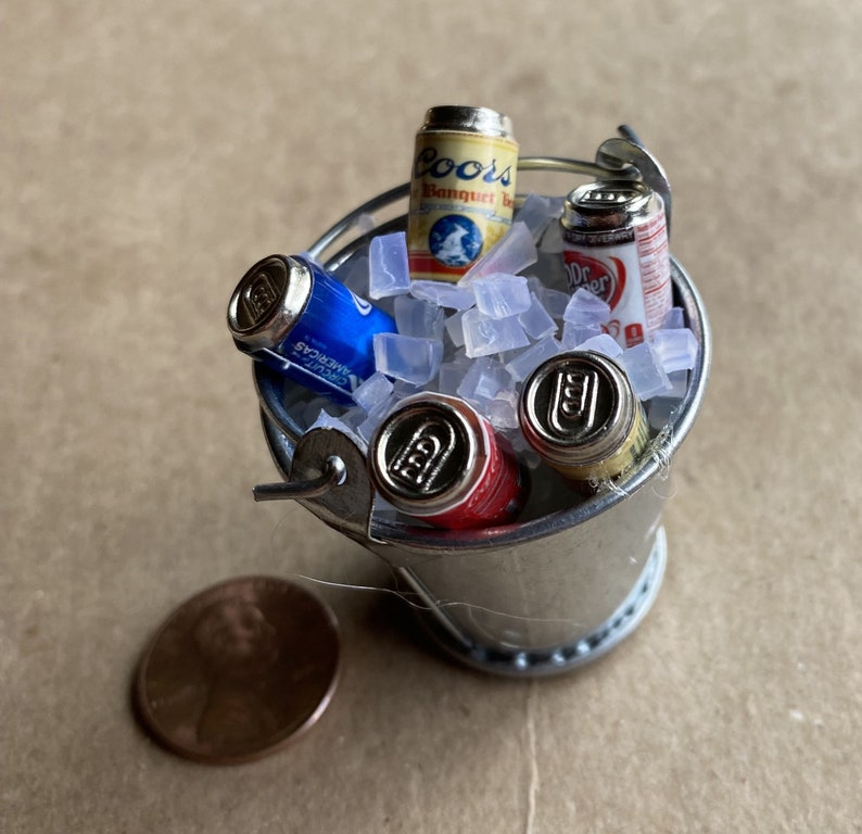 Puede incluir: Un cubo de metal plateado lleno de hielo y cuatro latas de cerveza en miniatura. Las latas son Coors Banquet Beer, Dr Pepper y otras dos marcas.