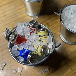 Puede incluir: Un cubo de metal plateado lleno de hielo y latas en miniatura de refrescos y cerveza. Las latas incluyen Dr Pepper, Coors Light y una lata azul con una etiqueta ilegible.