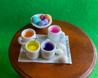 May include: Four small white mugs filled with different colored liquids, a bowl of pastel colored Easter eggs, and white eggs on a newspaper on a wooden table.