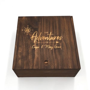 May include: A dark brown wooden box with a hinged lid. The lid is engraved with a compass, an arrow, and the text "The Adventures of Cooper & Mary Grace".