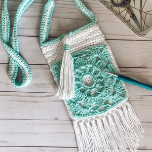 May include: A crocheted turquoise and white crossbody bag with a white tassel and fringe. The bag is on a white wooden surface.