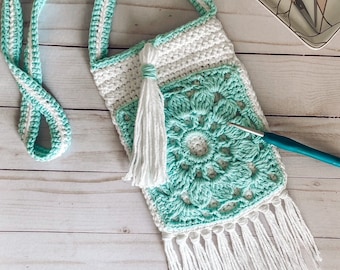 Puede incluir: Un bolso de hombro de crochet turquesa y blanco con un borla y flecos blancos. El bolso está sobre una superficie de madera blanca.