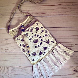 May include: A crocheted bag with a white floral design and brown accents. The bag has a long strap and a fringe trim.