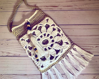 Puede incluir: Un bolso de crochet con un diseño floral blanco y detalles marrones. El bolso tiene una correa larga y un ribete de flecos.