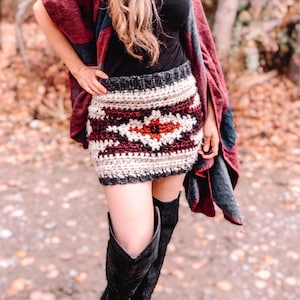 May include: A crocheted skirt with a geometric pattern in shades of brown, white, and red. The skirt is worn with black boots and a burgundy and blue plaid shawl.