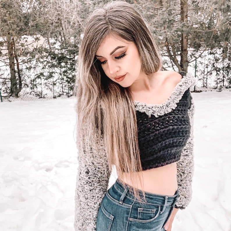 May include: A woman wearing a black and white cropped sweater with a fuzzy white trim. The sweater is worn over a pair of blue jeans.