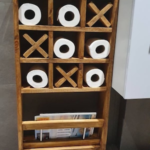 May include: A wooden bathroom storage shelf with a tic-tac-toe design. The shelf has three rows of compartments, each holding a roll of toilet paper. The bottom shelf has a magazine rack.