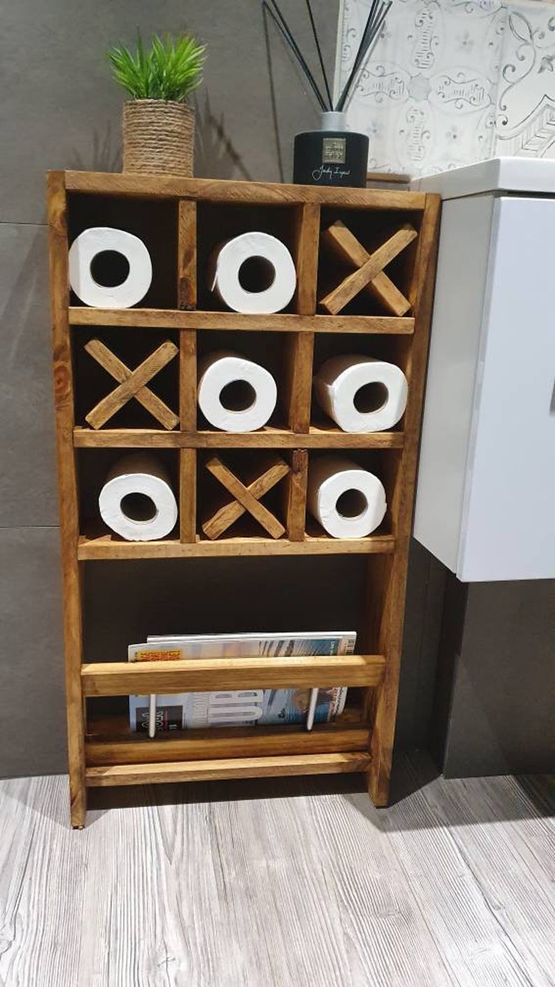 May include: A wooden bathroom storage shelf with a tic-tac-toe design. The shelf has three rows of compartments, each holding a roll of toilet paper. The bottom shelf has a magazine rack.