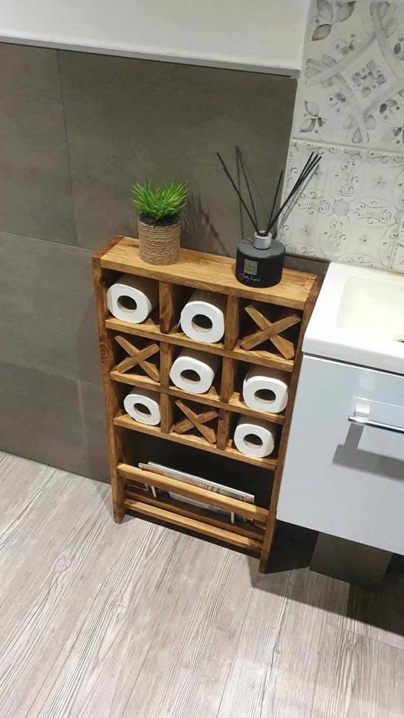 May include: A wooden toilet paper holder with a tic-tac-toe design. The holder has four shelves for storing toilet paper rolls and a lower shelf for storing magazines or other items. The holder is made of brown wood and has a natural finish.