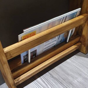 May include: A wooden shelf unit with two rolls of toilet paper and a magazine rack. The magazine rack has a magazine inside with the text "ACE" and "ONE DAY DELI".