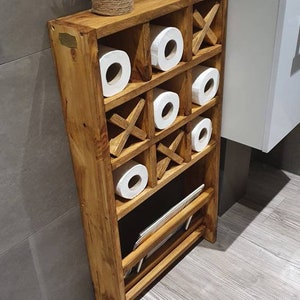 May include: A wooden bathroom storage shelf with a tic-tac-toe design. The shelf has six compartments for storing toilet paper rolls and a lower shelf for storing other items. The shelf is made of natural wood and has a rustic finish.