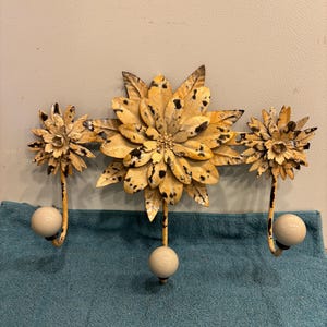 May include: A decorative wall hook with a floral design. The metal hook features three flower-shaped elements in yellow with black accents. Each hook has a cream-colored ball at the end. The wall hook is mounted on a light gray surface.