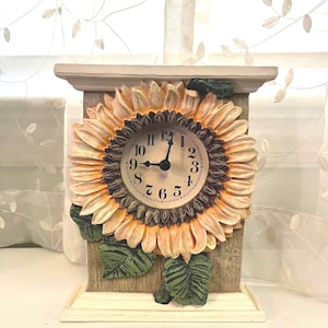 May include: A decorative clock featuring a sunflower design. The clock face is surrounded by a detailed sunflower with yellow petals and a brown center. The clock has a rectangular base and a cream-colored top.