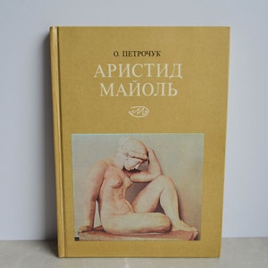Op de afbeelding: Een hardcover boek met een mosterdgele kaft met de titel "АРИСТИД МАЙОЛЬ" in witte Cyrillische letters. De kaft bevat een afbeelding van een naakt sculptuur en de naam van de auteur. Het boek staat rechtop.