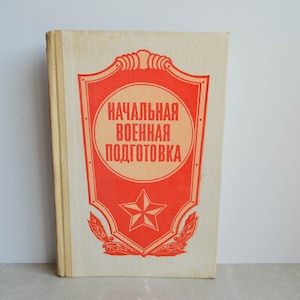 May include: A vintage book with a red shield on the cover. The shield has a red star and the text "НАЧАЛЬНАЯ ВОЕННАЯ ПОДГОТОВКА" in Russian.