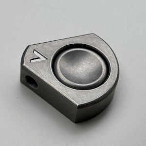 May include: A small, metallic gray fidget spinner with a rounded central button and a triangular design on one corner. The spinner has a distressed, textured finish and a small hole on one side.